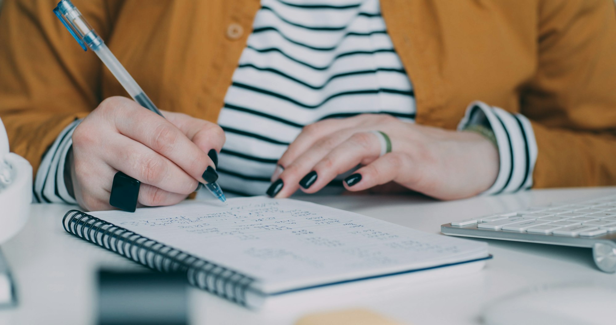 Hands writing figures into a spiral notebook beside a keyboard